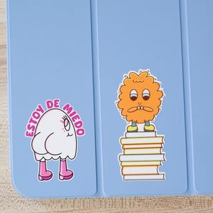 May include: A light blue tablet case with two cartoon stickers. One sticker features a white ghost-like character with pink boots and the text "Estoy de Miedo". The other sticker is an orange, fluffy character standing on books.