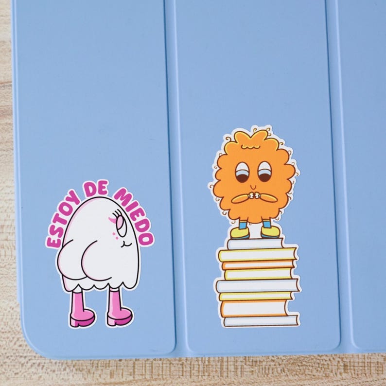 May include: A light blue tablet case with two cartoon stickers. One sticker features a white ghost-like character with pink boots and the text "Estoy de Miedo". The other sticker is an orange, fluffy character standing on books.