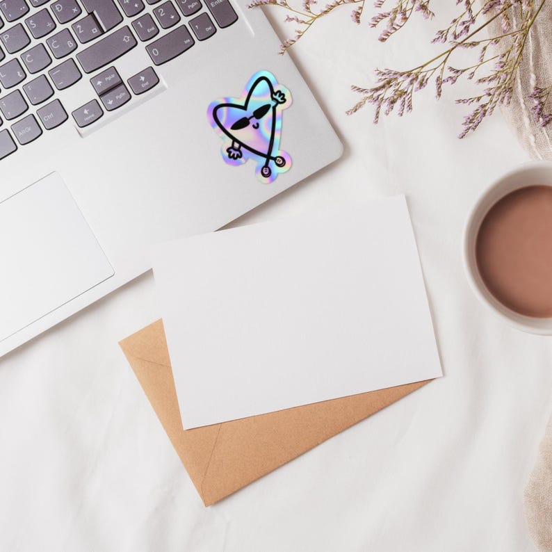 Puede incluir: Una pegatina hologr&aacute;fica de un coraz&oacute;n con gafas de sol y saludando, colocada en un port&aacute;til. Una tarjeta blanca en blanco y un sobre marr&oacute;n est&aacute;n sobre una superficie blanca, con una taza de l&iacute;quido marr&oacute;n y flores moradas.