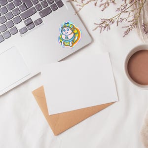 May include: A colorful sticker featuring a cartoon character in a blue and yellow outfit, with a matching hat and a yellow inner tube. The sticker is placed on a laptop, with a blank white card and a brown envelope nearby. A cup of coffee and some flowers are also in the image.