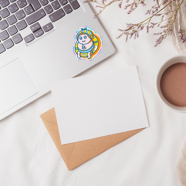 May include: A colorful sticker featuring a cartoon character in a blue and yellow outfit, with a matching hat and a yellow inner tube. The sticker is placed on a laptop, with a blank white card and a brown envelope nearby. A cup of coffee and some flowers are also in the image.
