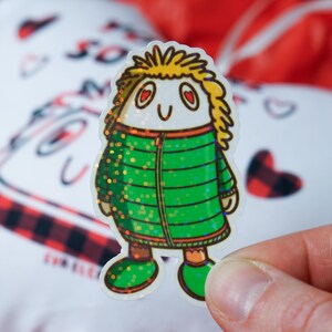 Puede incluir: Una pegatina con un personaje de dibujos animados con pelo amarillo, ojos en forma de coraz&oacute;n y una chaqueta verde con detalles brillantes. La pegatina se sostiene en una mano, con un fondo borroso de una almohada blanca en forma de coraz&oacute;n y tela roja.