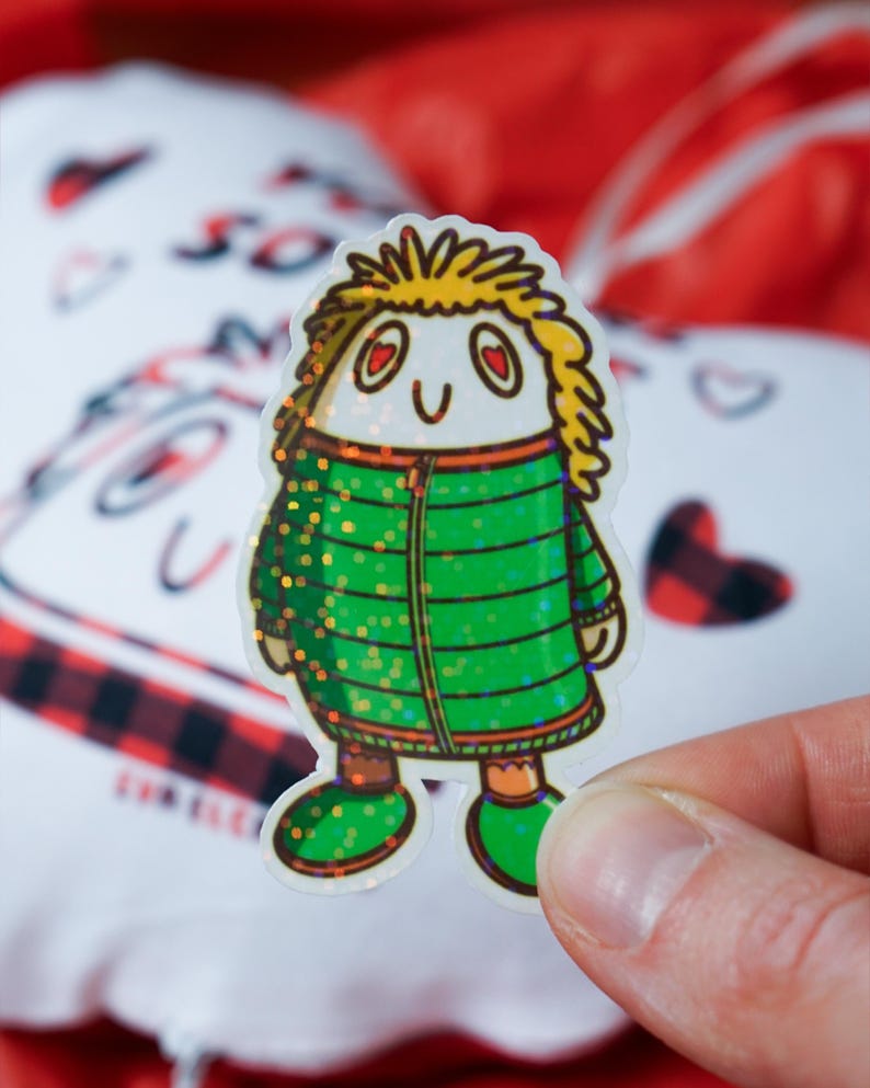 Puede incluir: Una pegatina con un personaje de dibujos animados con pelo amarillo, ojos en forma de coraz&oacute;n y una chaqueta verde con detalles brillantes. La pegatina se sostiene en una mano, con un fondo borroso de una almohada blanca en forma de coraz&oacute;n y tela roja.