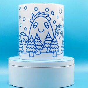 May include: White ceramic mug with a blue cartoon illustration of smiling mountain-like figures and trees. The design includes blue snowflakes. The mug is on a white rotating stand against a blue background.