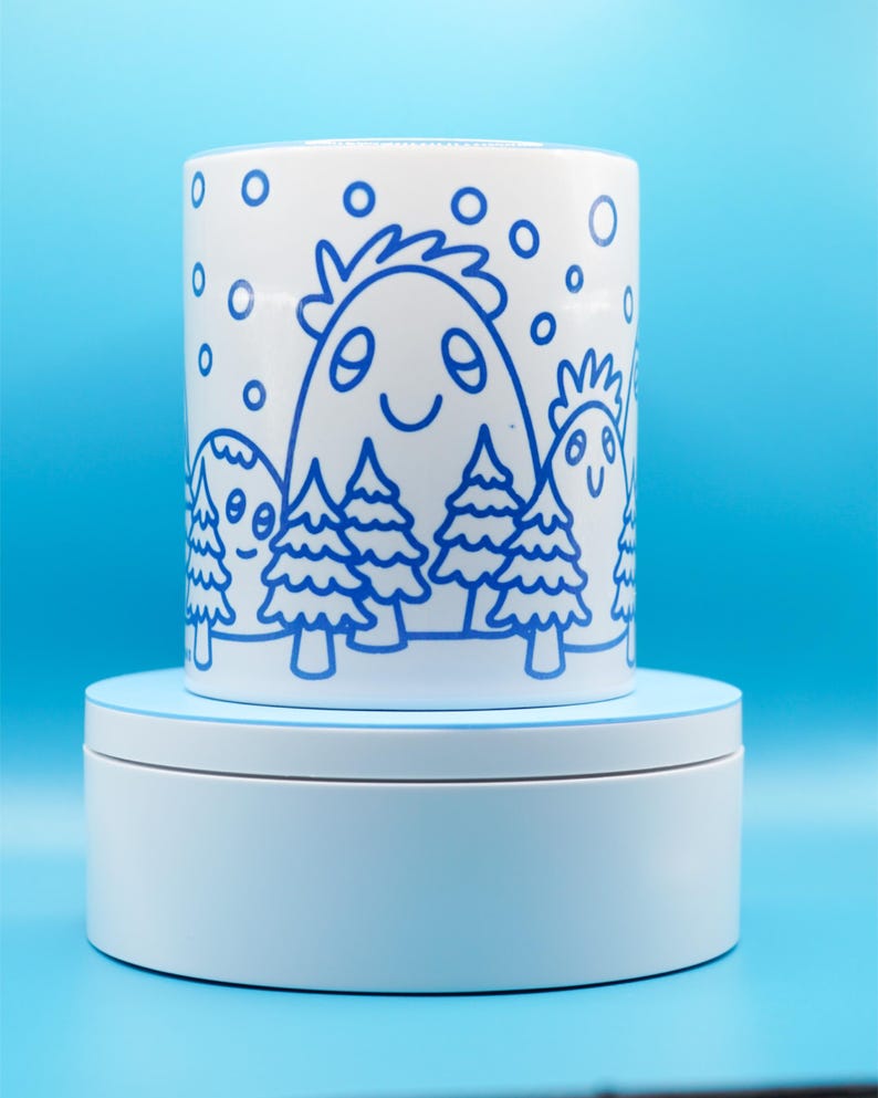 May include: White ceramic mug with a blue cartoon illustration of smiling mountain-like figures and trees. The design includes blue snowflakes. The mug is on a white rotating stand against a blue background.