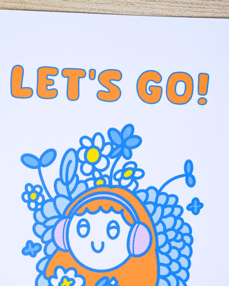 May include: A white print featuring the text "LET'S GO!" in orange. Below the text is a cartoon illustration of a person with orange hair wearing headphones, surrounded by blue flowers and leaves.