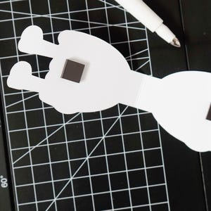 K&ouml;nnte beinhalten: Wei&szlig;er Papierauschnitt in Form einer Hand mit einem quadratischen Magneten. Ein wei&szlig;er Stift liegt auf einer schwarzen Schneidematte mit einem Rastermuster. Der Handausschnitt hat zwei ausgestreckte Finger.