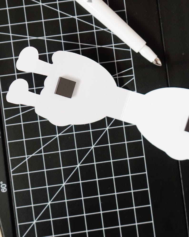 K&ouml;nnte beinhalten: Wei&szlig;er Papierauschnitt in Form einer Hand mit einem quadratischen Magneten. Ein wei&szlig;er Stift liegt auf einer schwarzen Schneidematte mit einem Rastermuster. Der Handausschnitt hat zwei ausgestreckte Finger.
