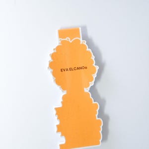 Op de afbeelding: Een oranje gestanste sticker met de tekst "EVA ELCANO" in een schreefloos lettertype. De sticker heeft een unieke vorm met een rechthoekige bovenkant en een gestileerde, wolkvormige onderkant. De sticker ligt op een wit oppervlak.