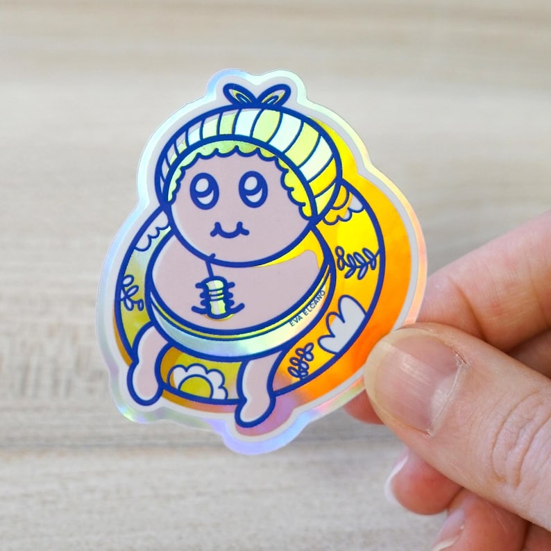 May include: Holographic sticker featuring a cartoon baby with a blue and yellow striped hat, a drink, and a yellow halo. The sticker has a rainbow sheen and is held by a hand. The sticker has the text "EVA ELCANO".