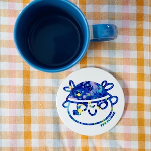 May include: A blue ceramic mug with a matching handle and a white coaster. The coaster features a cartoon face with a blue and yellow design and the text "EVA ELCANO". The mug and coaster are on a yellow and white checkered surface.