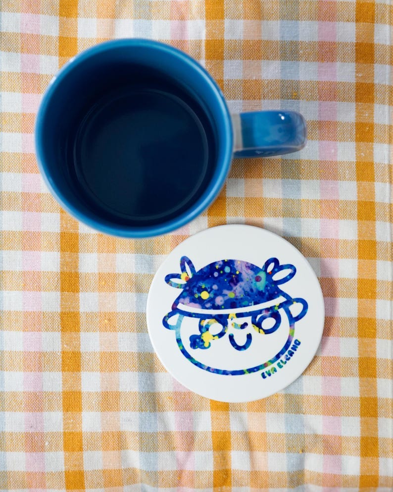 May include: A blue ceramic mug with a matching handle and a white coaster. The coaster features a cartoon face with a blue and yellow design and the text "EVA ELCANO". The mug and coaster are on a yellow and white checkered surface.