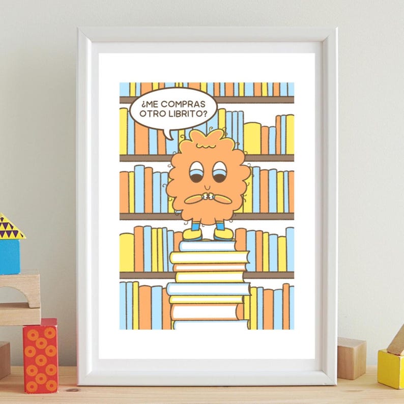 May include: A framed cartoon print featuring a whimsical orange character standing on a stack of books in front of a bookshelf. The character has a speech bubble with text in Spanish. The print has a white border and is displayed on a light-colored surface.