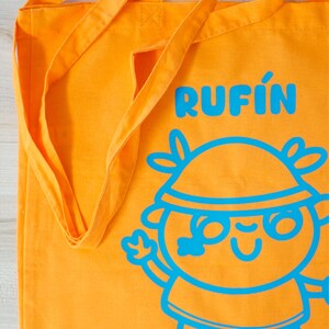 Może przedstawiać: Pomarańczowa torba typu tote z długimi paskami. Torba zawiera słowo "RUFIN" w kolorze niebieskim, wraz z ilustracją postaci z kresk&oacute;wki w kolorze niebieskim. Torba jest wykonana z wytrzymałej tkaniny.