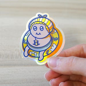 May include: Holographic sticker featuring a cartoon baby with a yellow and blue striped hat and a matching circular design. The baby has a serene expression and is surrounded by decorative floral elements. The sticker has a shiny, iridescent finish.