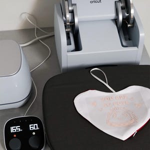 Puede incluir: Una m&aacute;quina de prensa de calor Cricut con una pantalla digital que muestra 165 grados Fahrenheit (74 grados Celsius) y 60 segundos. Un objeto blanco en forma de coraz&oacute;n con el texto "YOU ARE SO CUTE" est&aacute; sobre la superficie negra de la prensa.