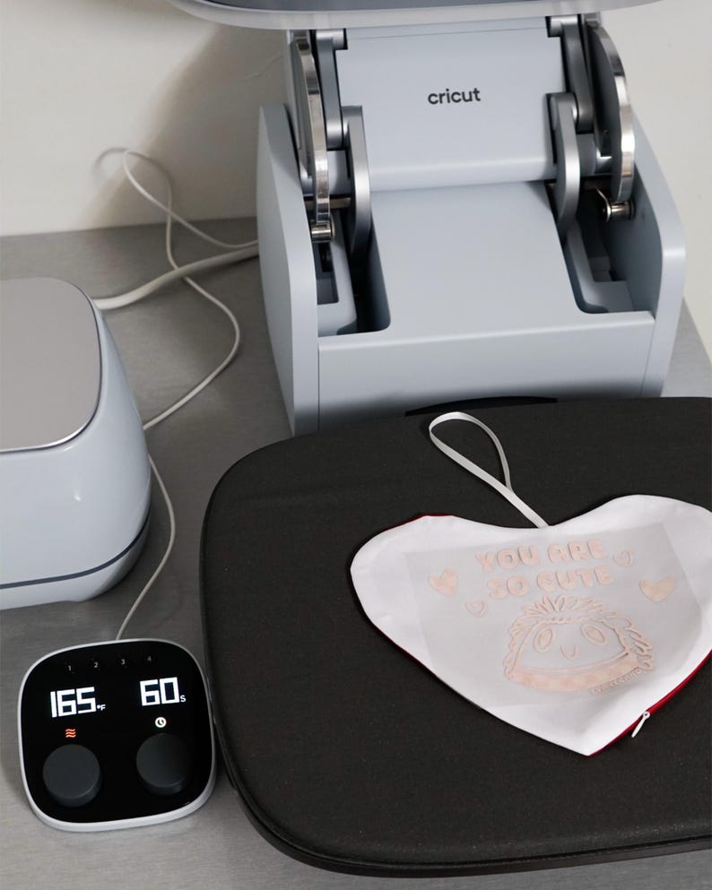 Puede incluir: Una m&aacute;quina de prensa de calor Cricut con una pantalla digital que muestra 165 grados Fahrenheit (74 grados Celsius) y 60 segundos. Un objeto blanco en forma de coraz&oacute;n con el texto "YOU ARE SO CUTE" est&aacute; sobre la superficie negra de la prensa.