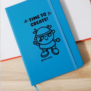 May include: A vibrant blue notebook featuring the words "TIME TO CREATE!" and a cartoon character. It has a blue elastic closure and a ribbon bookmark. The notebook is on a wooden surface. The notebook measures approximately 21 cm in length.