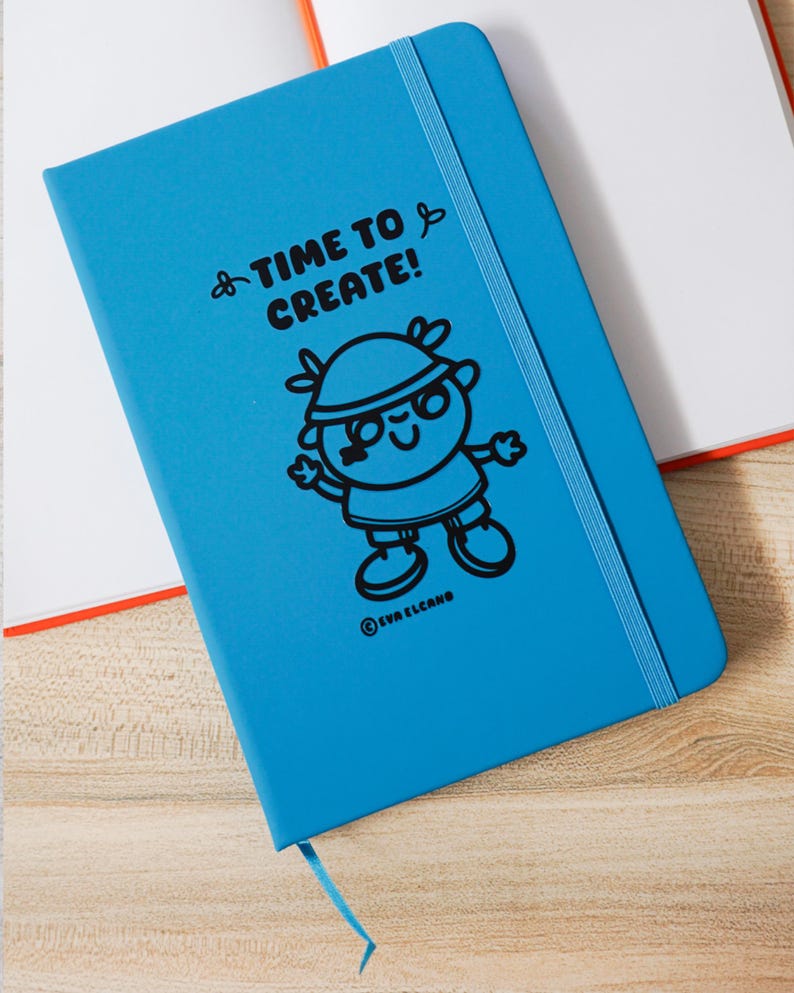 May include: A vibrant blue notebook featuring the words "TIME TO CREATE!" and a cartoon character. It has a blue elastic closure and a ribbon bookmark. The notebook is on a wooden surface. The notebook measures approximately 21 cm in length.