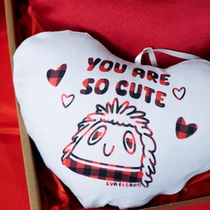 Puede incluir: Una almohada blanca en forma de coraz&oacute;n con el texto "YOU ARE SO CUTE" en letras rojas y negras. La almohada presenta un personaje de dibujos animados con un dise&ntilde;o de cuadros rojos y negros y peque&ntilde;os corazones.