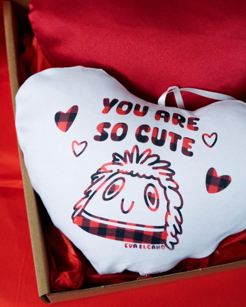 Puede incluir: Una almohada blanca en forma de coraz&oacute;n con el texto "YOU ARE SO CUTE" en letras rojas y negras. La almohada presenta un personaje de dibujos animados con un dise&ntilde;o de cuadros rojos y negros y peque&ntilde;os corazones.