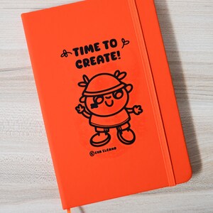 May include: An orange notebook featuring the text "TIME TO CREATE!" and a cartoon character illustration. The notebook has an elastic band closure and a ribbon bookmark. The artist's name, EVA ELCANO, is printed at the bottom.