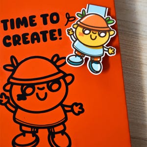 May include: An orange notebook featuring the text "TIME TO CREATE!" and a cartoon illustration. A sticker of a cartoon character with an orange hat, blue shirt, and glasses is attached. The illustration is in black.