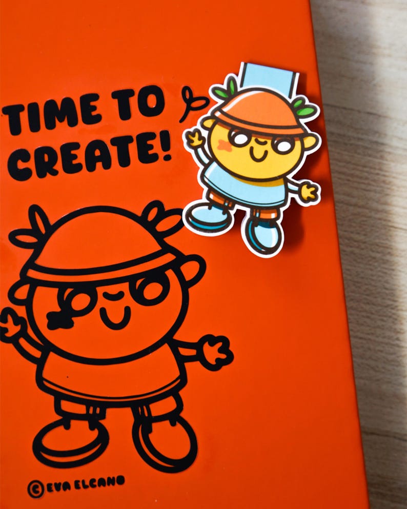 May include: An orange notebook featuring the text "TIME TO CREATE!" and a cartoon illustration. A sticker of a cartoon character with an orange hat, blue shirt, and glasses is attached. The illustration is in black.