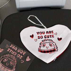 Puede incluir: Un adorno de tela blanco en forma de coraz&oacute;n con el texto "YOU ARE SO CUTE" y un personaje de dibujos animados con un dise&ntilde;o de cuadros rojos y negros. El adorno tiene un lazo blanco para colgar y ribete rojo.