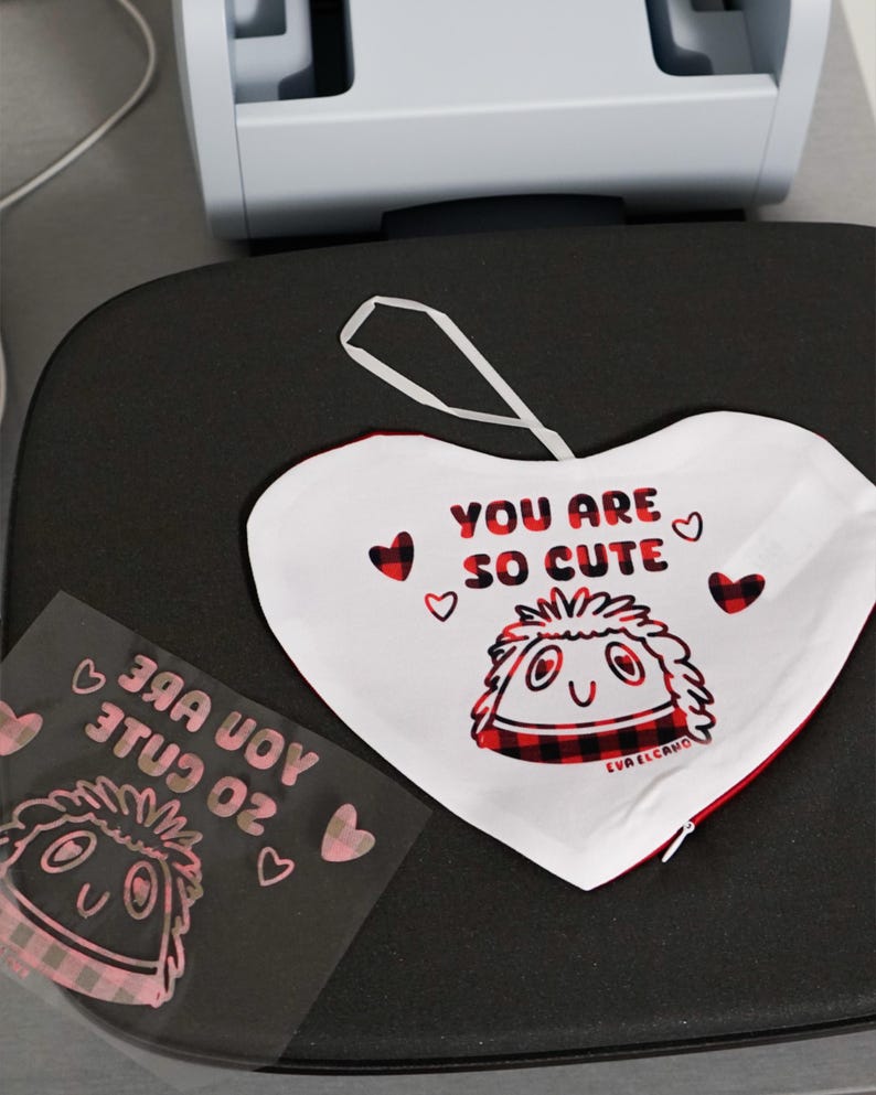 Puede incluir: Un adorno de tela blanco en forma de coraz&oacute;n con el texto "YOU ARE SO CUTE" y un personaje de dibujos animados con un dise&ntilde;o de cuadros rojos y negros. El adorno tiene un lazo blanco para colgar y ribete rojo.