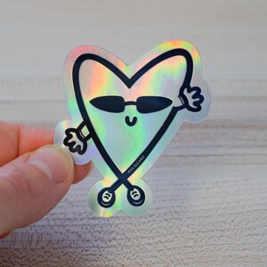 Puede incluir: Pegatina hologr&aacute;fica con un personaje en forma de coraz&oacute;n con gafas de sol y brazos. El personaje tiene una cara sonriente y est&aacute; delineado en negro. La pegatina tiene un acabado brillante e iridiscente. El texto "EVA ELCANO" est&aacute; impreso en la parte inferior.