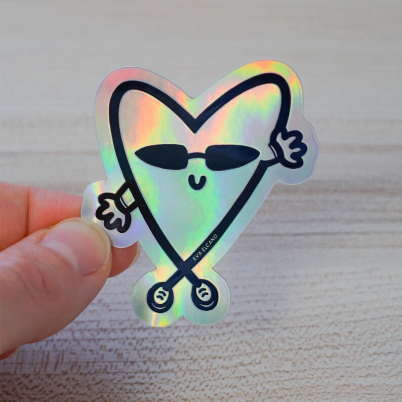 Puede incluir: Pegatina hologr&aacute;fica con un personaje en forma de coraz&oacute;n con gafas de sol y brazos. El personaje tiene una cara sonriente y est&aacute; delineado en negro. La pegatina tiene un acabado brillante e iridiscente. El texto "EVA ELCANO" est&aacute; impreso en la parte inferior.