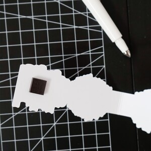 Op de afbeelding: Een witte papieren uitsparing met een donkerbruin vierkantje bevestigd, rustend op een zwarte snijmat met een wit rasterpatroon. Een witte pen ligt diagonaal bovenop.