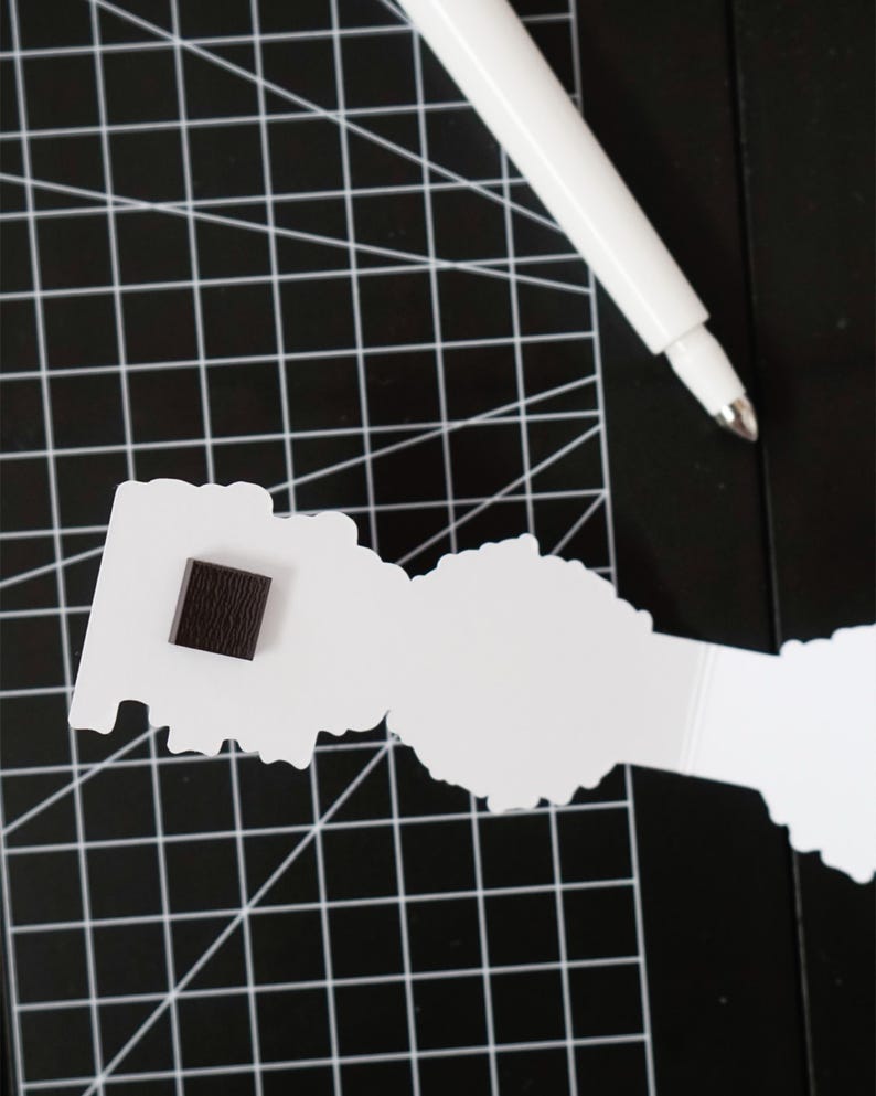 Op de afbeelding: Een witte papieren uitsparing met een donkerbruin vierkantje bevestigd, rustend op een zwarte snijmat met een wit rasterpatroon. Een witte pen ligt diagonaal bovenop.
