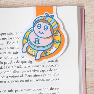 May include: A colorful bookmark featuring a cartoon baby design. The bookmark has an orange tab and a circular image of a baby with a blue and yellow striped hat, surrounded by a yellow floral design. The baby is pink and blue.
