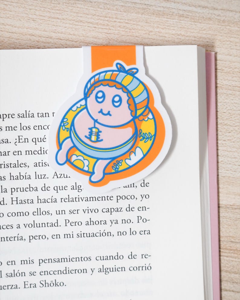 May include: A colorful bookmark featuring a cartoon baby design. The bookmark has an orange tab and a circular image of a baby with a blue and yellow striped hat, surrounded by a yellow floral design. The baby is pink and blue.