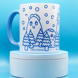 May include: White ceramic mug with a blue handle and a whimsical blue illustration of mountains, trees, and falling snow. The mug features the text "EVA EL CANO" at the bottom. The mug is sitting on a light blue stand.