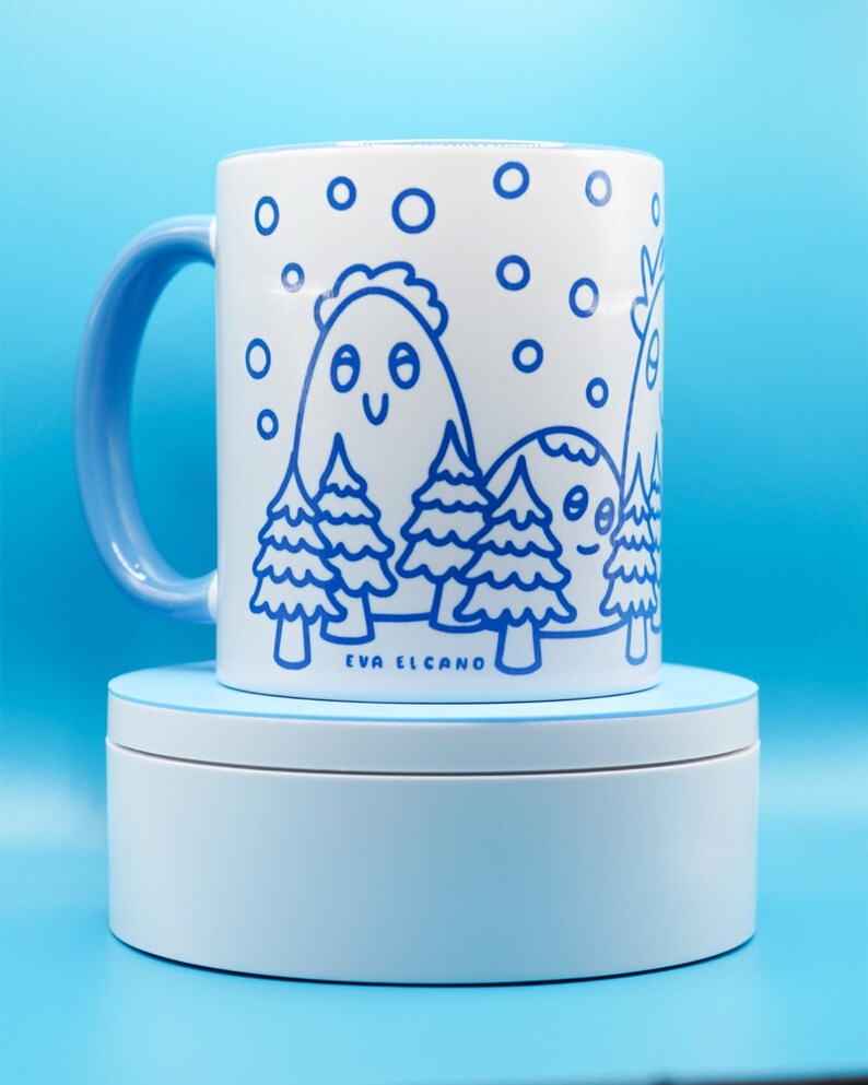 May include: White ceramic mug with a blue handle and a whimsical blue illustration of mountains, trees, and falling snow. The mug features the text "EVA EL CANO" at the bottom. The mug is sitting on a light blue stand.