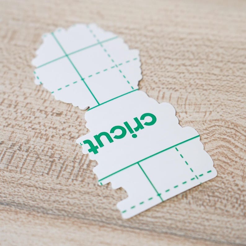 May include: A white Cricut cutting mat with a green grid pattern and the brand name "Cricut" printed in green. The mat has a unique, irregular shape and is designed for crafting projects.