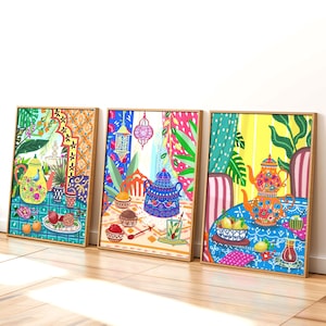 May include: Three framed art prints featuring colorful illustrations of tea settings. Each print showcases a different vibrant scene with teapots, fruits, and decorative elements. The artwork is set against a white wall and wooden floor.