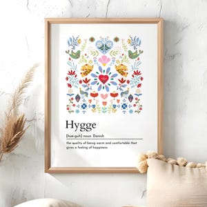 May include: A framed print with a vibrant floral and bird pattern, the word "Hygge", and its definition. The design incorporates red, blue, yellow, and green hues. The frame is a light brown colour, adding a natural touch to the artwork.
