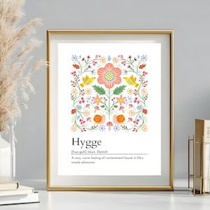 Può includere: Una stampa artistica incorniciata con una cornice dorata presenta la parola "Hygge" e un colorato design floreale con uccelli. È inclusa anche la definizione di Hygge. L'opera d'arte è esposta su una superficie bianca.