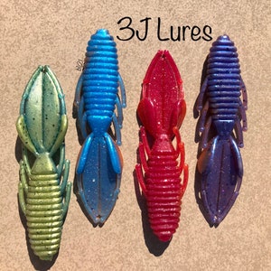 May include: Four plastic fishing lures in different colors: green, blue, red, and purple. The lures are shaped like crawfish and have a segmented body. The text "3J Lures" is written above the lures.