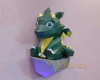 May include: A green and yellow papercraft dragon sitting on a purple rock. The dragon has large blue eyes and a yellow underbelly.