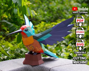 May include: A colorful papercraft hummingbird model with blue, green, orange, and purple accents. The model is perched on a brown papercraft base. The image includes text: "YouTube VIDEO TUTORIAL", "PDF", "STUDIO3", "SVG", "COMPATIBLE WITH CUTTING MACHINE".