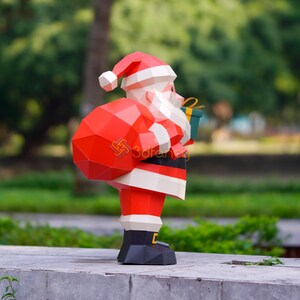 May include: A geometric papercraft Santa Claus figure, featuring a red suit, hat, and sack, with white trim and beard. He holds a green gift box with a yellow ribbon. The figure is standing on a concrete surface with a blurred green background.