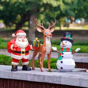 May include: A festive Christmas display featuring papercraft figures of Santa Claus, a reindeer, and a snowman. Santa wears a red suit and carries a sack. The reindeer holds a gift box. The snowman has a top hat and scarf.