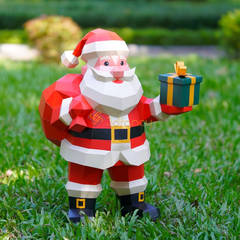 May include: A papercraft Santa Claus figure, wearing a red suit with white trim, a black belt with a gold buckle, and black boots. He holds a green gift box with a gold ribbon. The figure is standing on green grass.