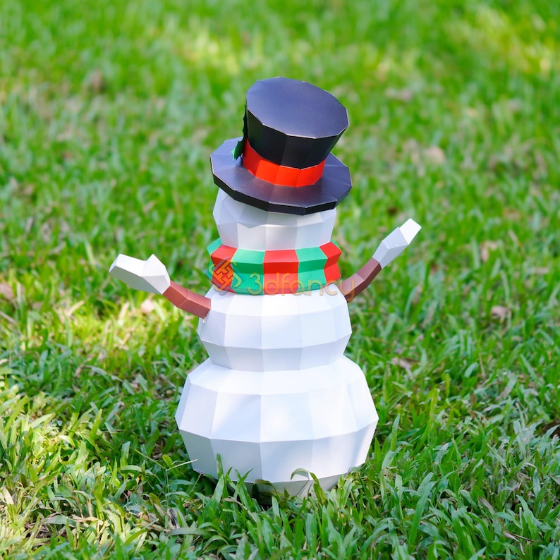 May include: A white, geometric paper snowman with a black top hat, a red and green scarf, and brown arms. The snowman is standing in green grass, with its arms outstretched. A festive winter decoration.