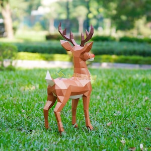 May include: A brown geometric papercraft reindeer with a red nose and large antlers stands on green grass. The reindeer is made of many triangular and polygonal shapes. The background is a blurred green park setting.
